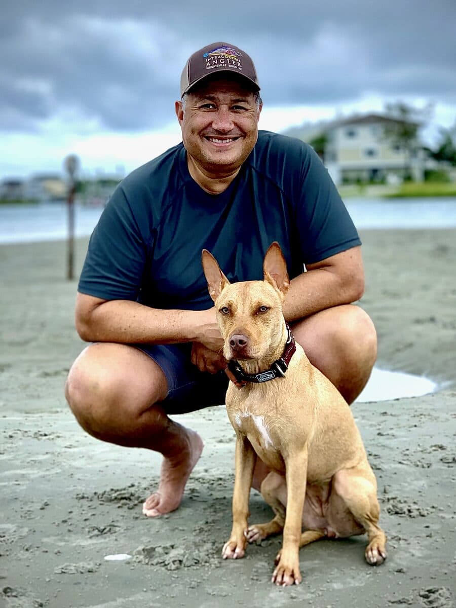 Ralph Sierra crouching on the sand at Wrightsville Beach with his dog